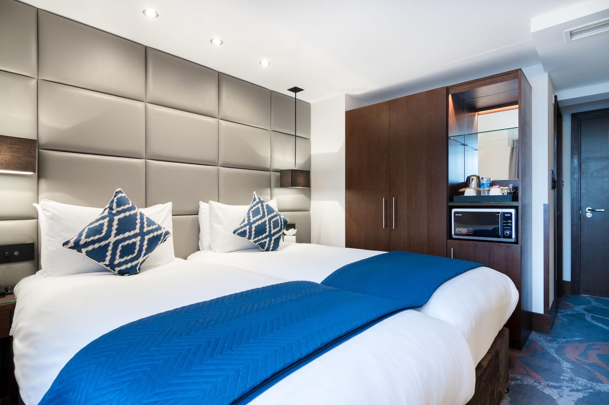 Montcalm Chilworth Townhouse, Paddington Hotel Londres Exterior foto The photo shows a modern hotel room featuring two beds with white linens and decorative blue throw blankets. Each bed has a pair of patterned cushions. The walls behind the beds are adorned with a sleek, padded design. To the side, there’s a wooden c