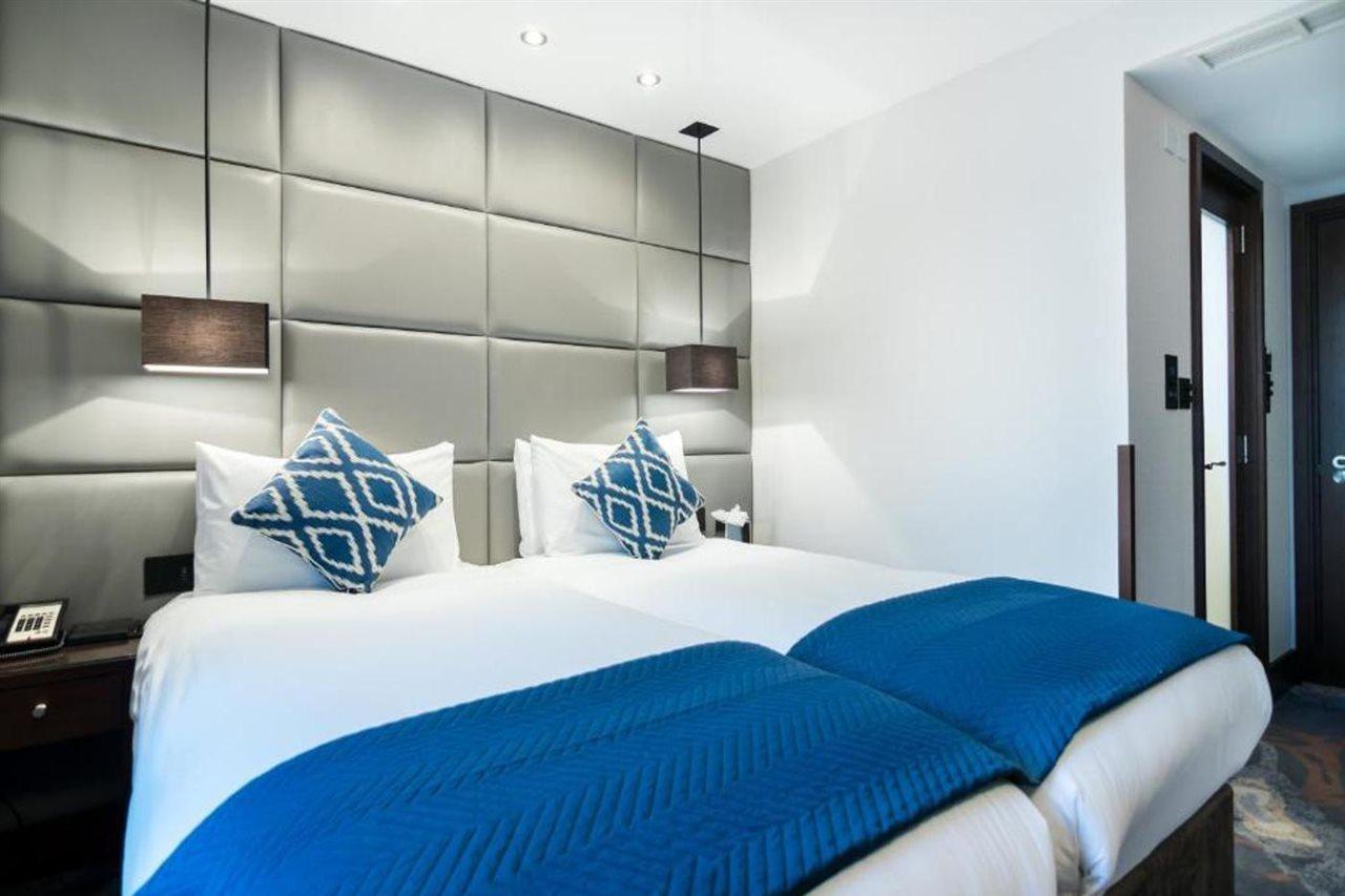 Montcalm Chilworth Townhouse, Paddington Hotel Londres Exterior foto The image shows a hotel room with two twin beds positioned close together. Each bed is adorned with white linens and a blue throw blanket. The pillows are white with decorative blue and white patterned pillows. Behind the beds is a stylish gray padde