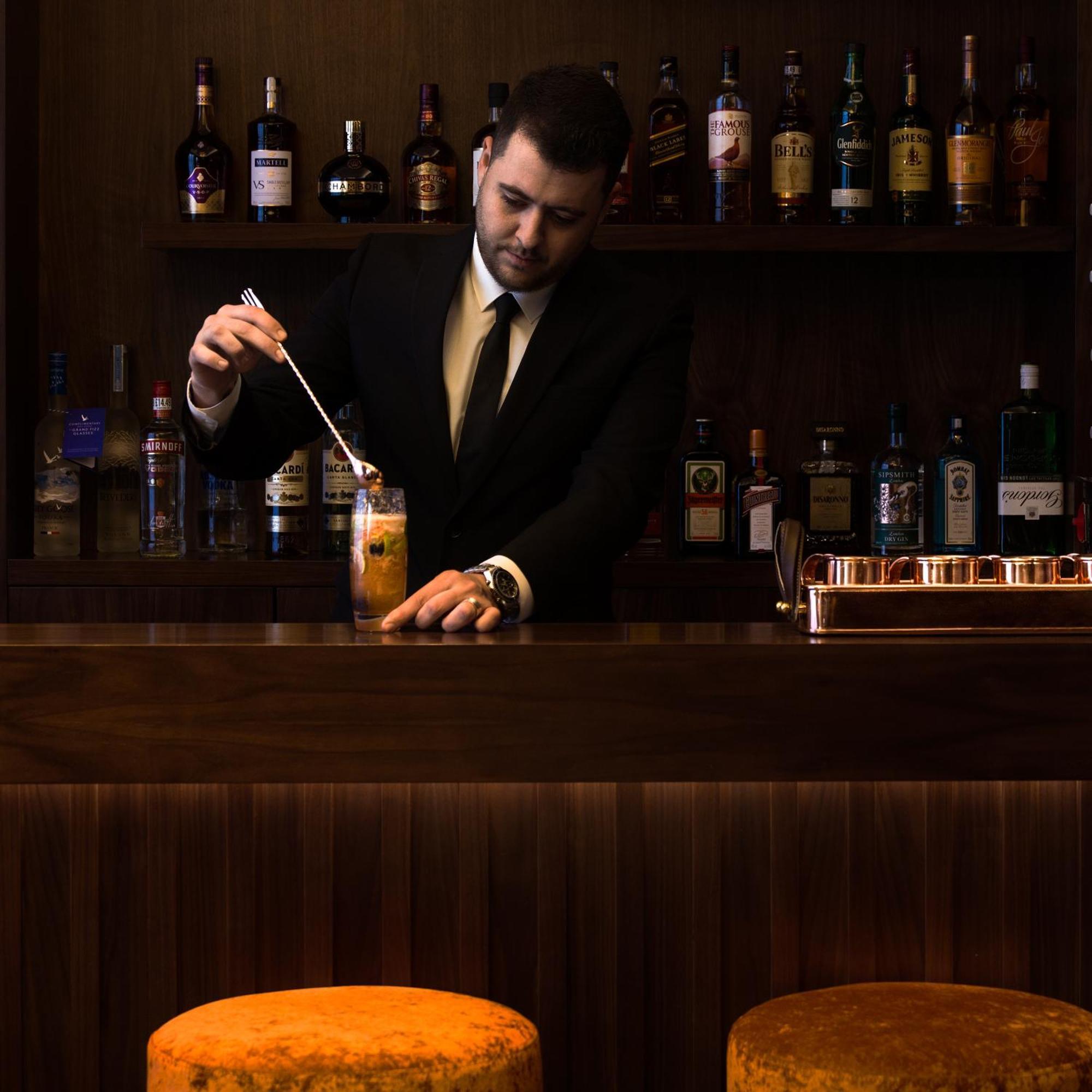 Montcalm Chilworth Townhouse, Paddington Hotel Londres Exterior foto The image shows a bartender at a bar. He is wearing a black suit and tie and is focused on preparing a cocktail, stirring it with a spoon. Behind him, there is a wooden bar counter and a shelf filled with various bottles of liquor and spirits. The at